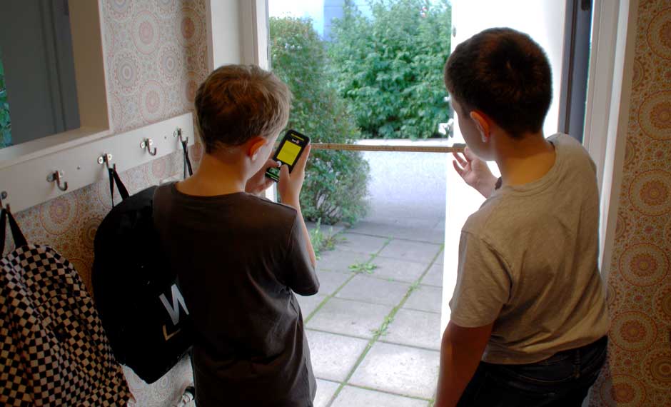 Två barn mäter en dörröppning med tumstock och svarar på frågor i en mobiltelefon.